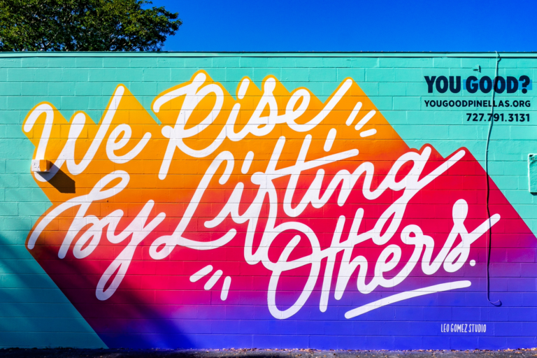 Colorful hand lettered mural by Leo Gomez saying "We Rise by Lifting Others"